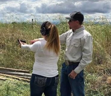 A man instructs a woman on how to shoot a handgun outdoors.