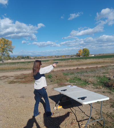 A woman shooting a handgun outdoors at a shooting range on a sunny day.