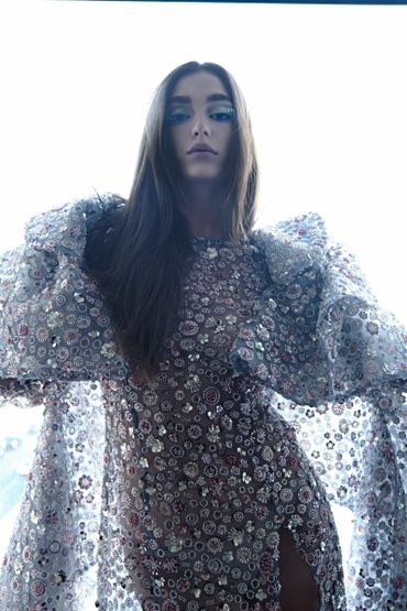 Woman in an intricately embellished, sheer gown with long hair against a bright background.