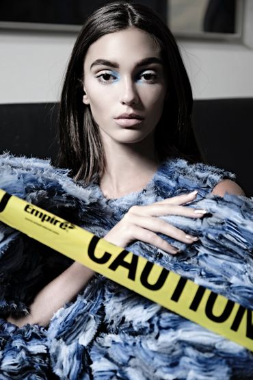 Woman with blue makeup behind yellow caution tape wearing a textured blue garment.