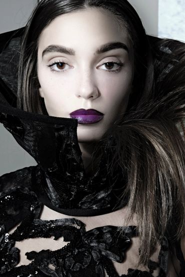 Close-up of a woman with bold purple lipstick and a high black collar garment.