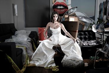 Woman in an elegant white dress amidst a cluttered, chaotic room with caution tape.