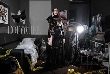 Model in a sheer black dress adorned with butterflies, posing in a cluttered room with caution tape.