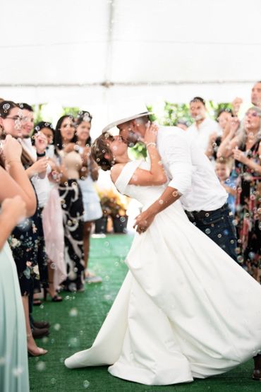Groom dips bride for a kiss in an aisle of friends and family as they all blow sparkling bubbles.