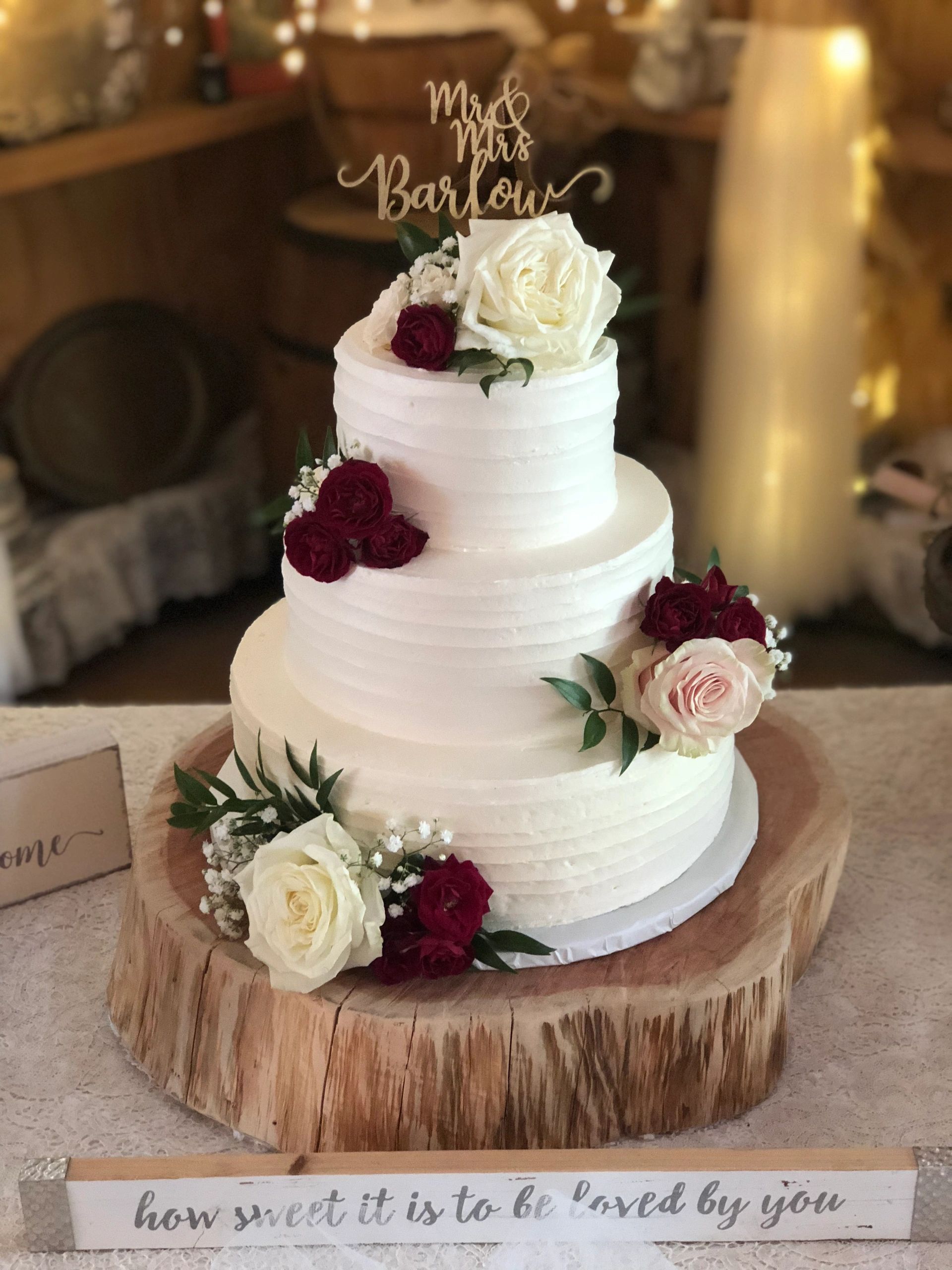 Three-tiered white wedding cake with roses and a wooden sign.