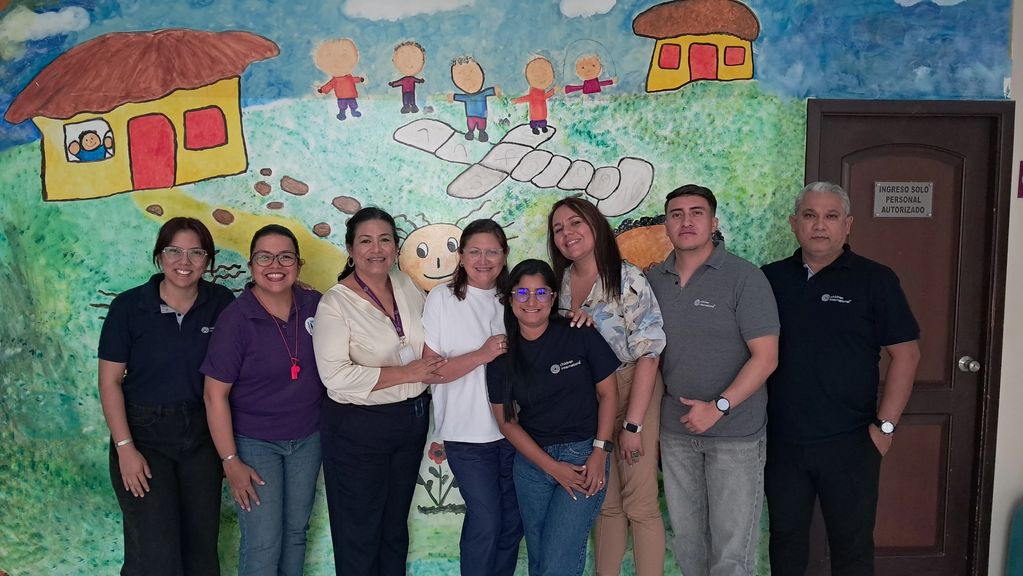 Group photo of eight smiling adults standing in front of a colorful mural with houses and children.