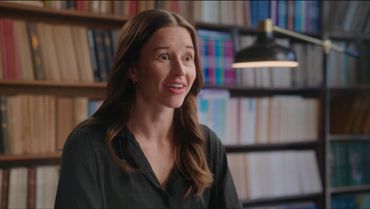A woman speaking in a library setting with bookshelves behind her.