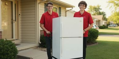 Two young men in red shirts moving a white refrigerator outside a house.