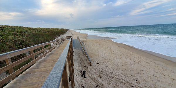 Wooden boardwalk leading to a sandy beach with ocean waves under a cloudy sky.