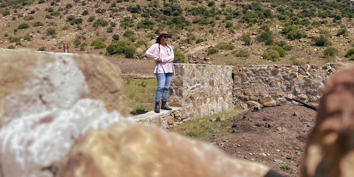 Person in a hat stands on a stone wall in a rocky, grassy landscape.