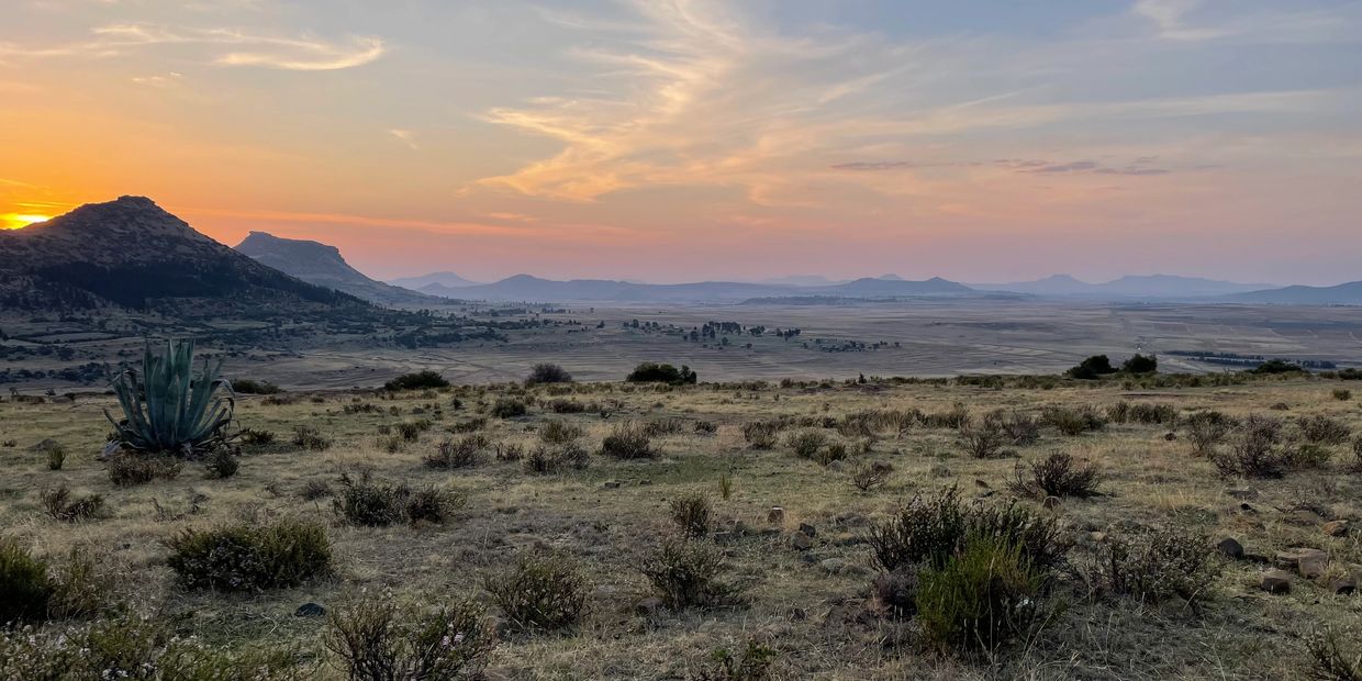 Sunset over a vast, dry landscape with distant mountains and scattered bushes.