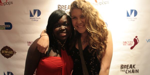 Two young women smiling and posing together in front of a promotional backdrop.