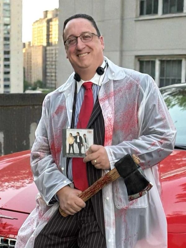 Man in bloody raincoat holding axe and CD, smiling.