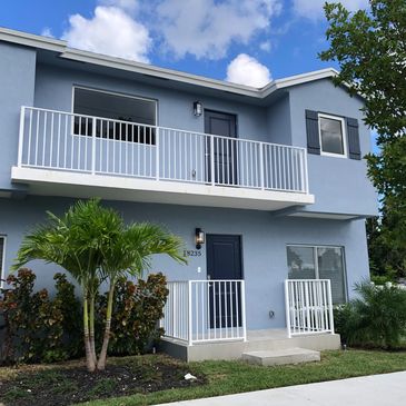 Blue two-story house with white railings and lush greenery.