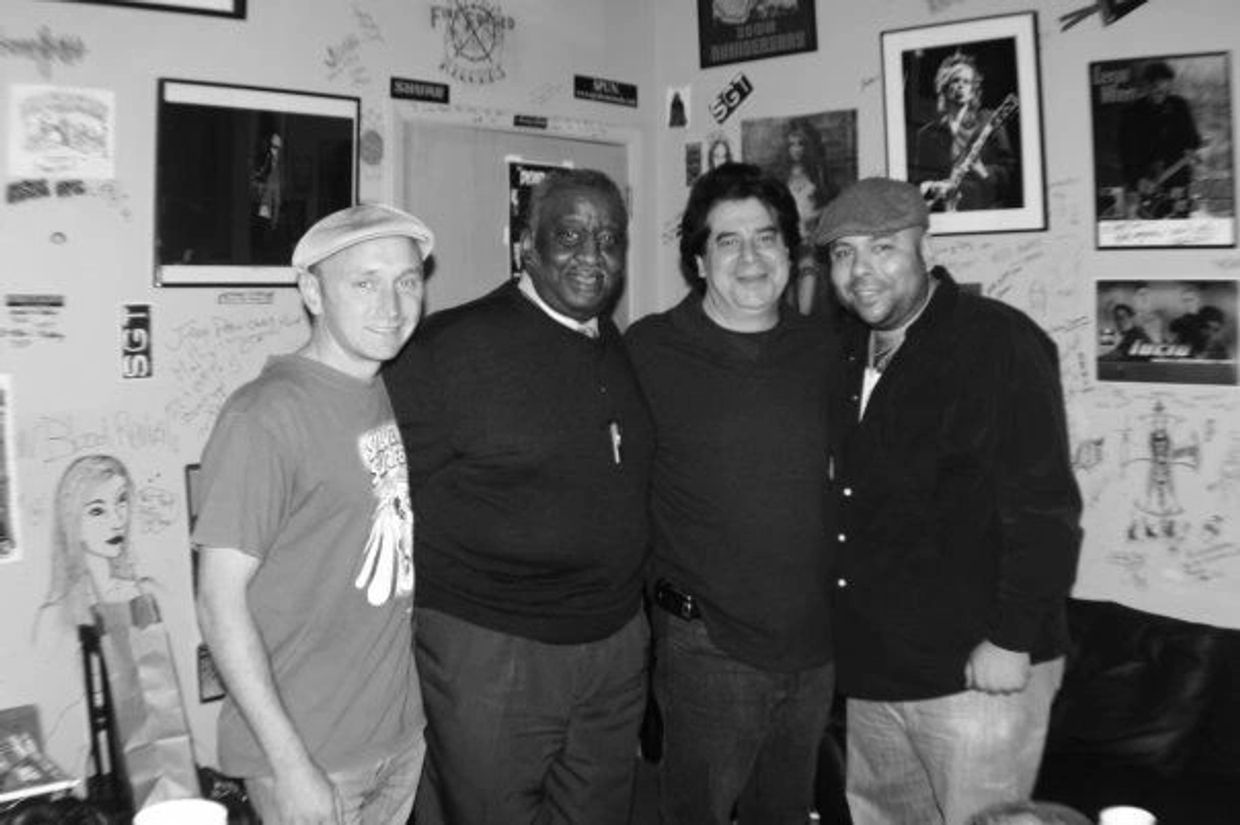 Four men posing together in a room with music posters.