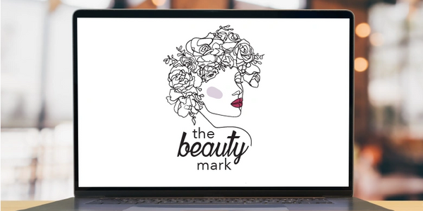 Laptop on desk with a line drawn logo of a lady with flowers in her hair, words the beauty mark.