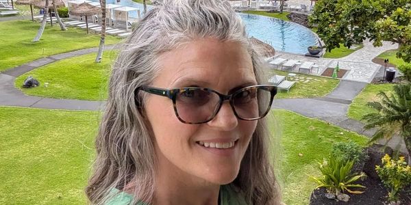 Smiling woman with glasses outdoors near a pool in a green garden.