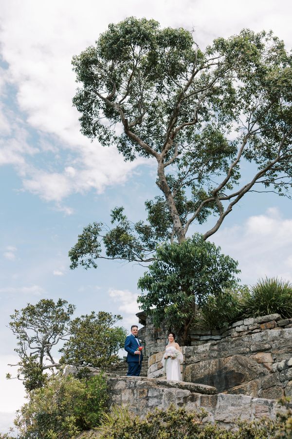 A couple in wedding attire stands on stone steps surrounded by greenery and trees under a blue sky.