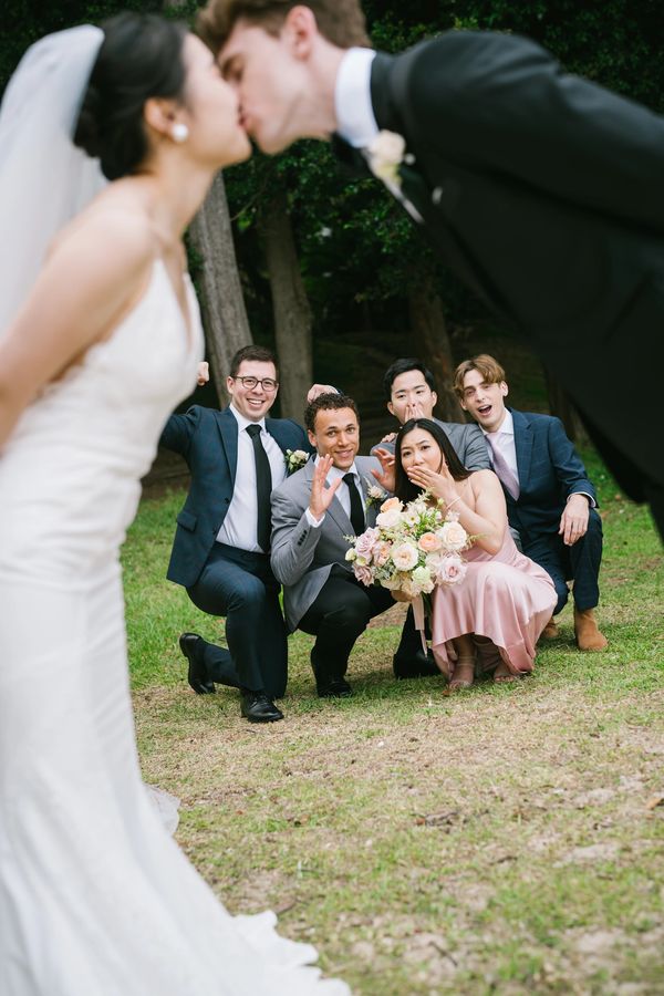 Bride and groom kiss in foreground with excited wedding guests reacting behind them.