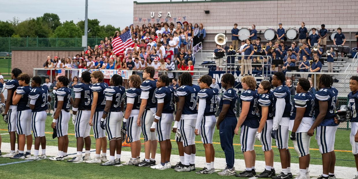 High school football players stand for the national anthem with cheering crowd and band behind.