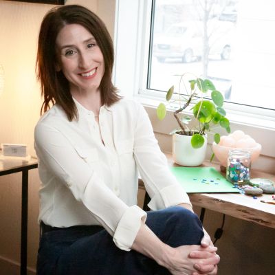 Smiling woman seated by a window with a plant and craft supplies on the table.