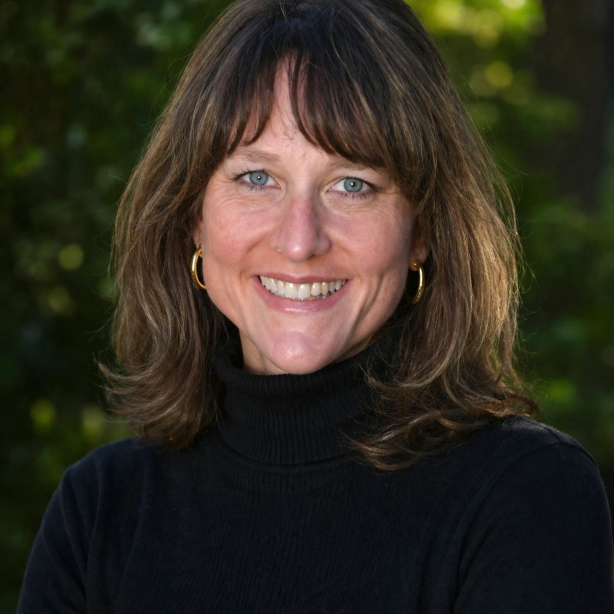 Smiling woman with brown hair wearing a black turtleneck outdoors.