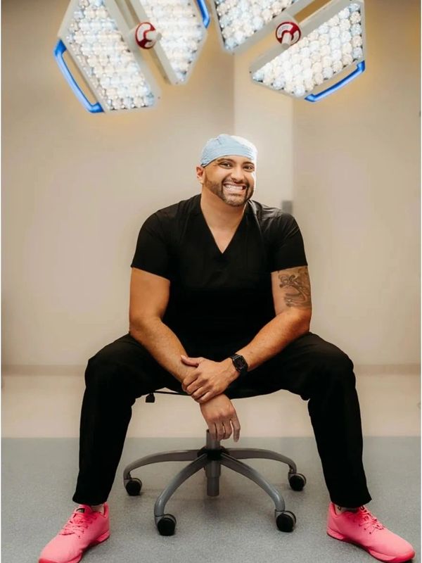 Smiling man in black scrubs with pink shoes sitting on a chair.