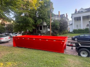 Red roll-off dumpster on a lawn beside trees and a house.