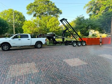 White pickup truck and roll-off trailer with the loading frame raised beside a red dumpster.