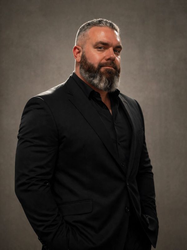 Man with a beard in a black suit posing confidently.