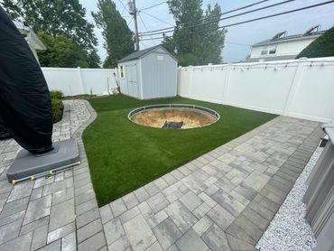 Backyard with a circular pit surrounded by artificial grass and stone pathways.