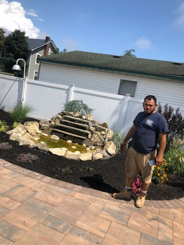 Man standing beside a landscaped garden with a small rock water feature.