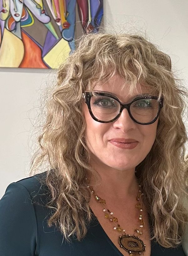 Woman with curly hair and glasses wearing a necklace and dark top.