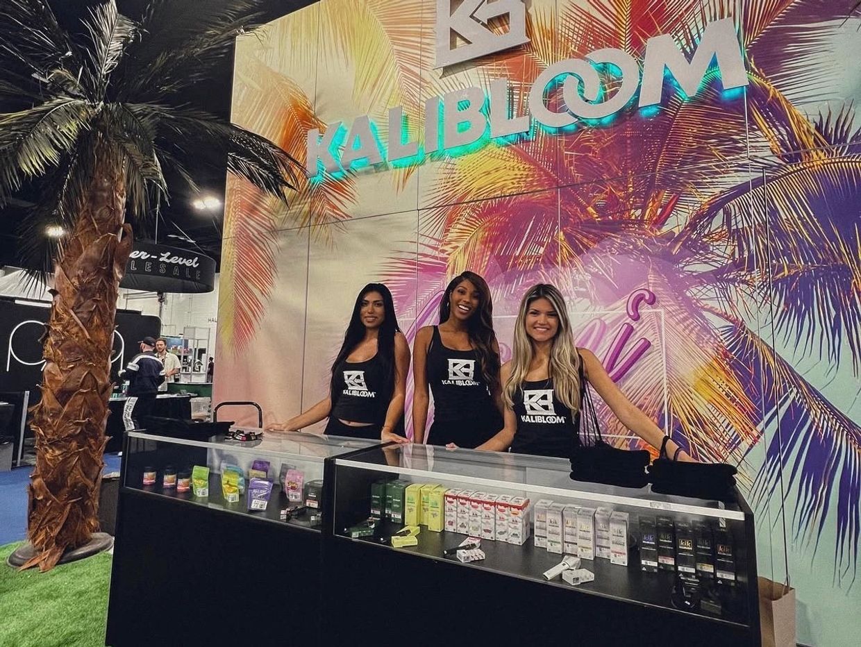 Three women at a Kalibloom promotional booth with tropical decor.