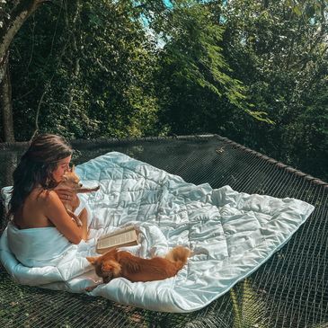 Woman cuddling cats on a hammock with a book in a forest setting.