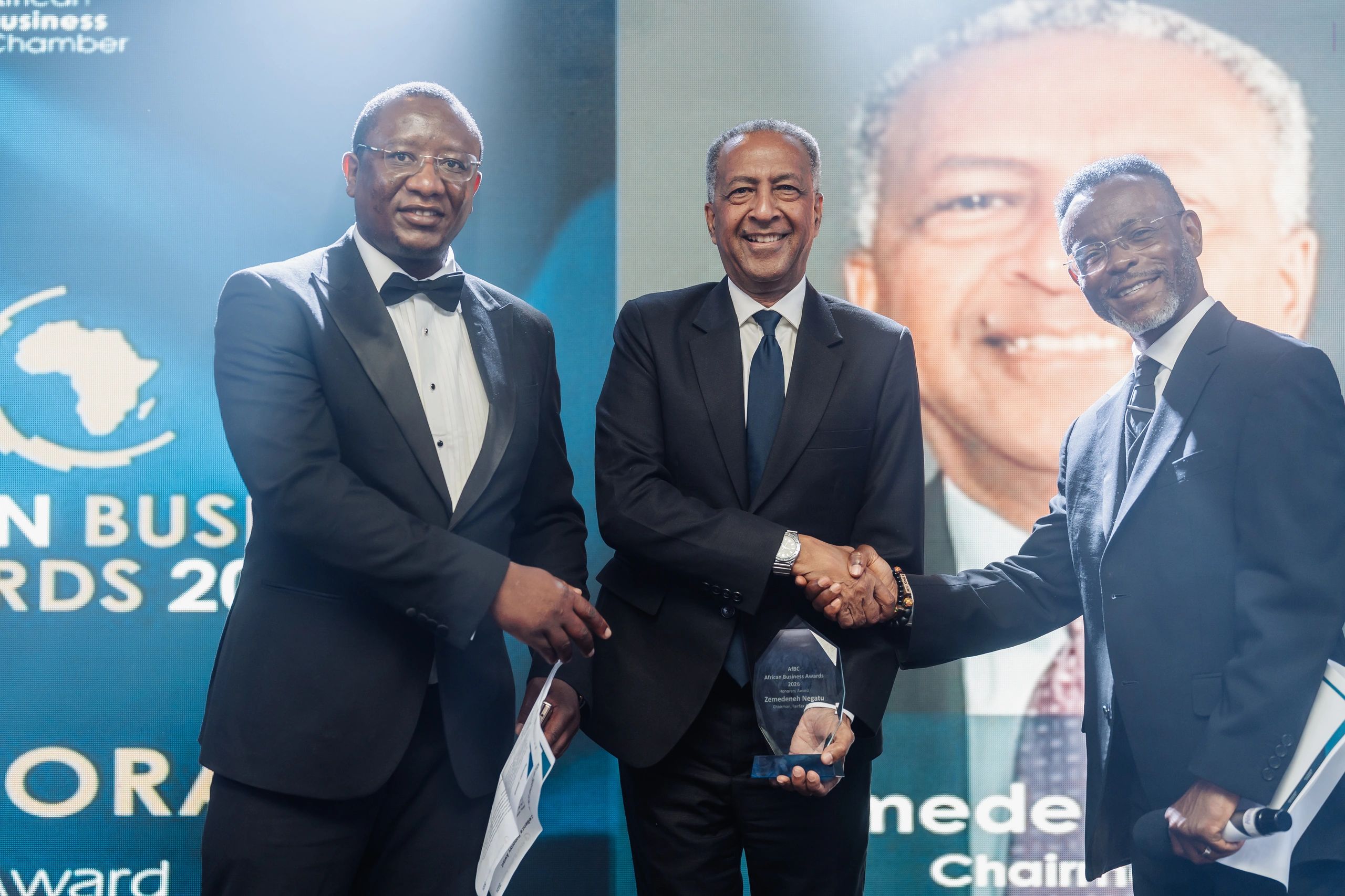 Three men in formal attire shaking hands at an award event.