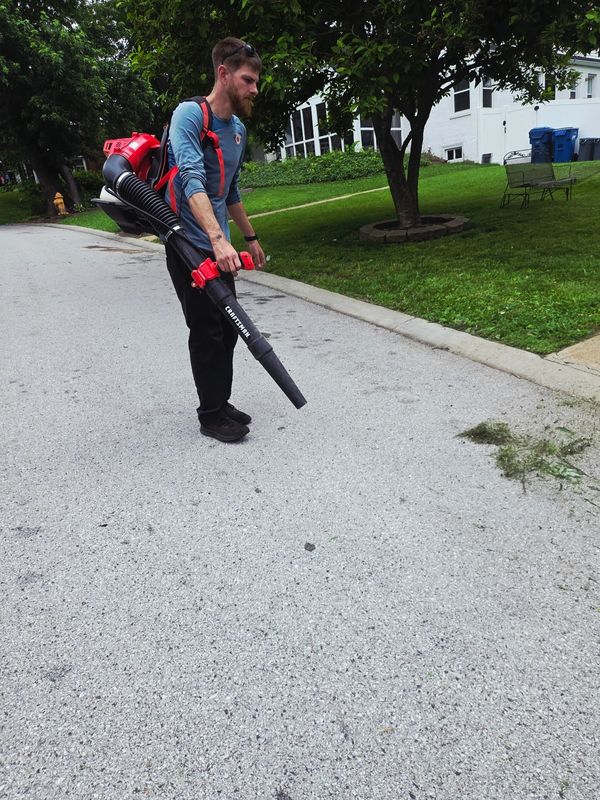 Man using a leaf blower to clear grass clippings on a driveway.