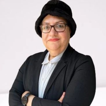 Confident woman with glasses and a black hat, dressed in a black blazer and white shirt.