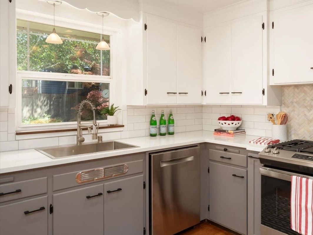 Modern kitchen with white and gray cabinets, stainless steel appliances, and a large window.