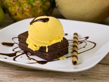 Brownie topped with yellow ice cream and chocolate drizzle on a white plate.