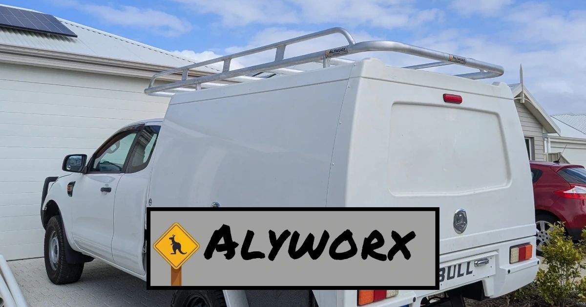 White utility truck with Alyworx canopy and roof rack parked in front of a house.