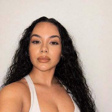 Woman with long curly hair and natural makeup poses against a plain wall.