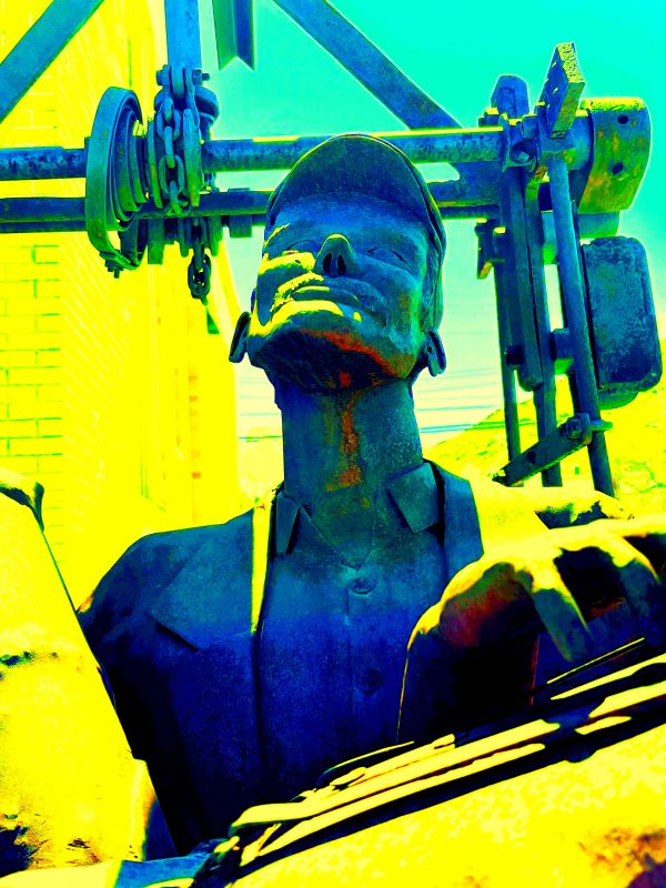 Metal statue of a man with a cap against industrial machinery.