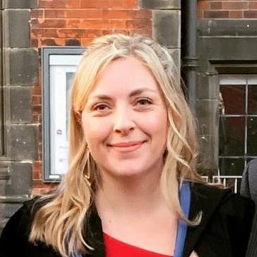 A smiling woman wearing a red dress and black coat with a blue and black lanyard.