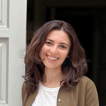 Smiling woman with wavy brown hair and casual outfit leaning against a white door.