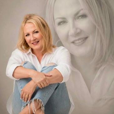 Smiling woman in casual white shirt and ripped jeans sitting with arms around knees.