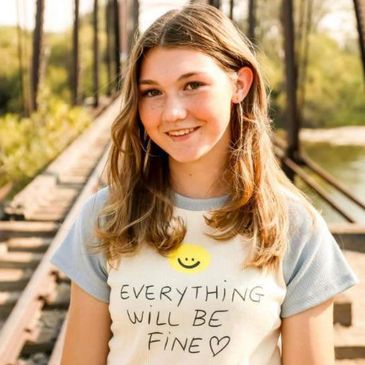 A smiling girl wearing a shirt that says 'Everything will be fine' on a sunny day.