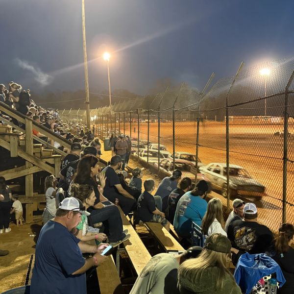 Crowd watching a dirt track car race at night.