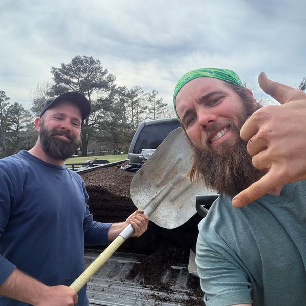 Two men with beards posing with a shovel and soil.