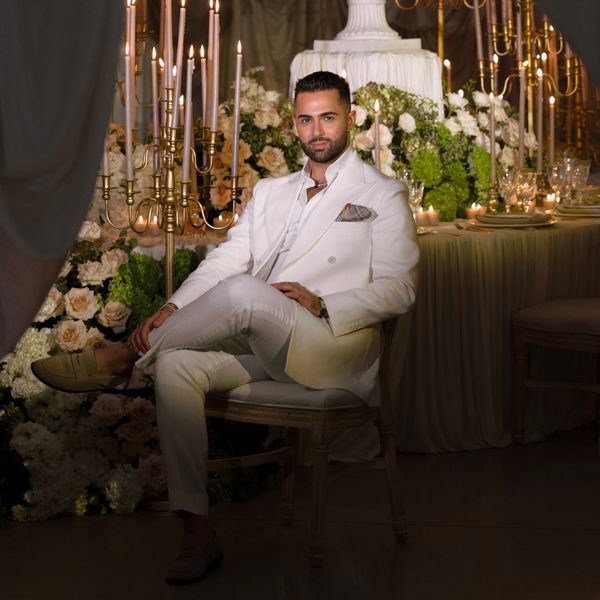 Man in an elegant white suit seated amid a lavish floral and candlelit setting.